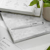 A silver metallic ballpoint pen resting on a white marble notepad, styled with an open recipe book, a potted green plant, and a beige ceramic pot on a white surface.